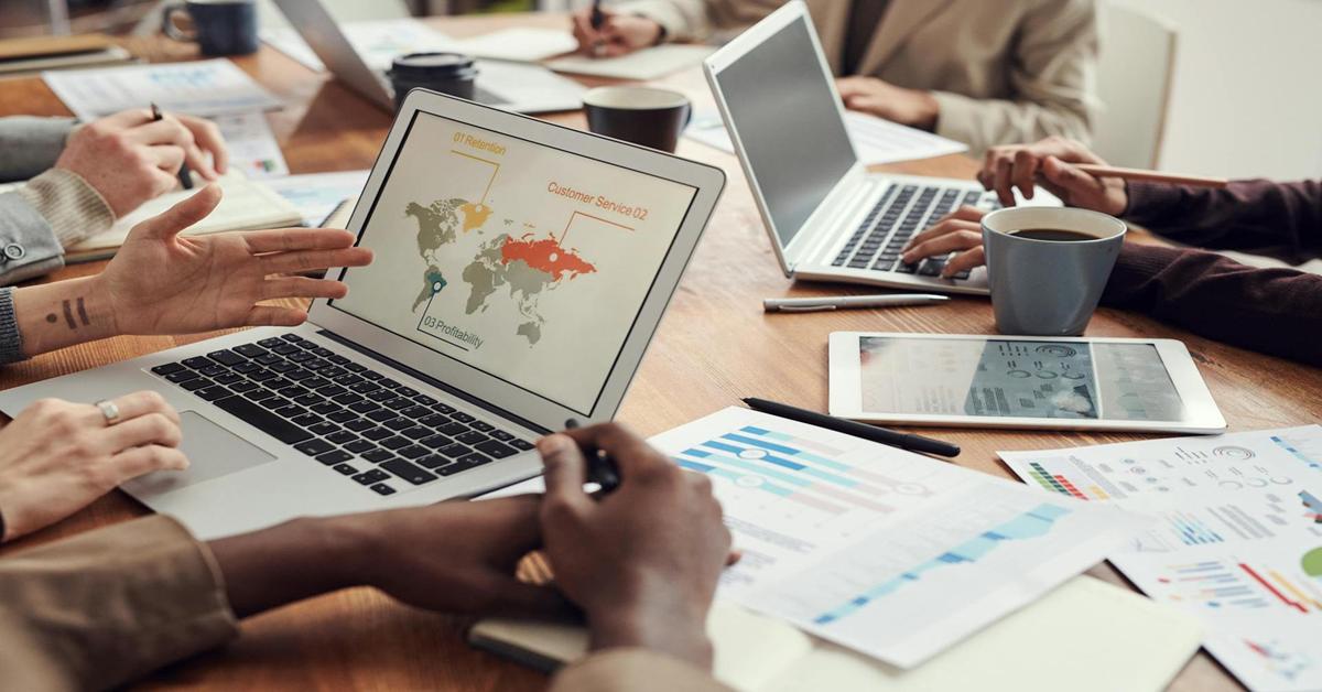 Group of professionals in a collaborative meeting around a wooden table with laptops, tablets, printed charts, and coffee cups, discussing Q2 global customer service results and marketing support strategies for small businesses.